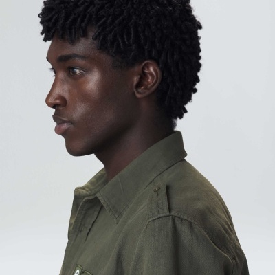 Perfil de jovem com camisa verde-oliva e patches, cabelo de caracóis grossos