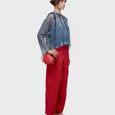 Mulher com blusa transparente azul, calças vermelhas largas, sandálias bege e bolsa vermelha