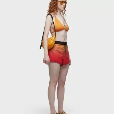 Mulher com bikini laranja, calções vermelhos e laranja, óculos de sol castanhos, bolsa amarela e sandálias douradas