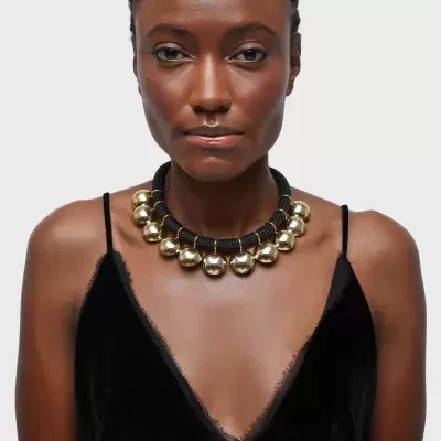 Colar com esferas douradas e base preta trançada no pescoço de mulher com vestido preto