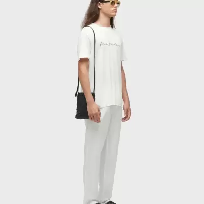 Pessoa com t-shirt branca com texto, calças brancas, sandálias pretas e óculos de sol