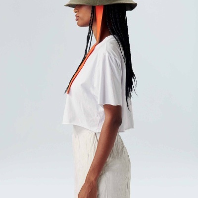 Mulher de perfil com chapéu bucket verde e cordão laranja, t-shirt branca e saia clara