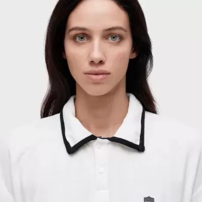 Polo branco com gola branca e borda preta, pequeno emblema em forma de escudo cinzento no peito esquerdo