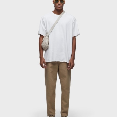 Homem com t-shirt branca, calças castanhas, sapatilhas bege, bolsa pequena bege ao ombro e óculos escuros amarelos