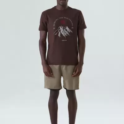 Homem com t-shirt castanha estampada, calções castanho claro e sapatilhas bege em fundo branco