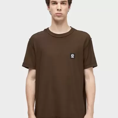 Homem a usar t-shirt castanha com etiqueta preta e letra e branca no peito