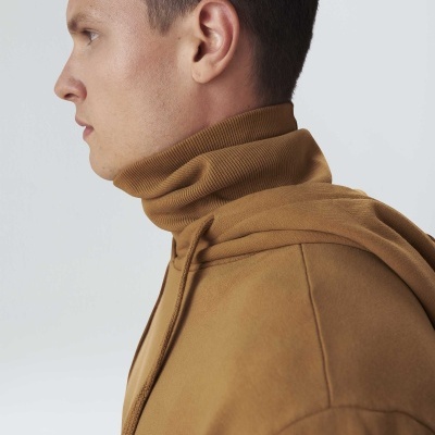 Homem com camisola de gola alta castanha e casaco castanho claro, perfil, fundo branco e cinza