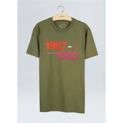 T-shirt verde com texto '1967 - 1969' em vermelho e rosa pendurada num cabide de madeira
