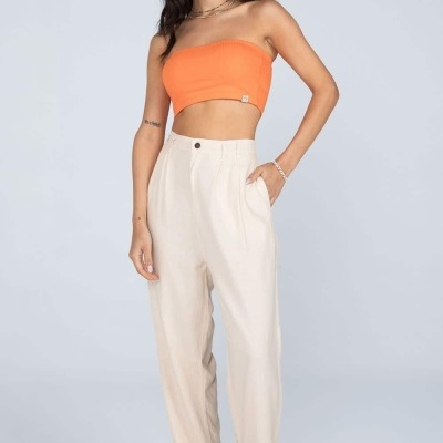 Mulher de pé com top laranja e calças bege claras. 