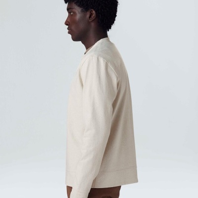 Homem de perfil com camisola bege e calças castanhas num fundo branco