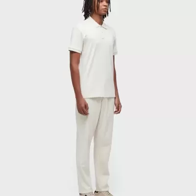 Homem vestido com camisola polo branca, calças brancas e ténis bege em fundo branco neutro