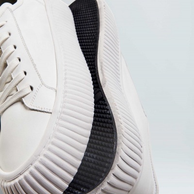 Sapatilhas brancas com sola espessa canelada e detalhe preto na sola sobre fundo cinza