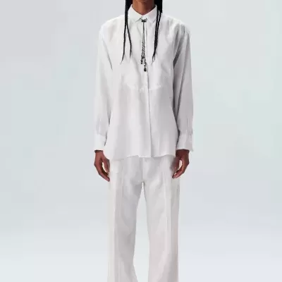 Pessoa vestindo camisa e calças brancas, colar preto, sapatos castanhos, fundo branco