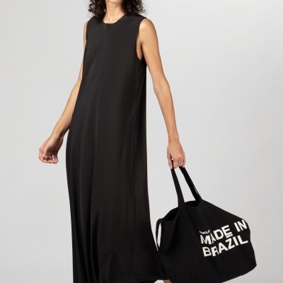 Mulher com vestido preto longo sem mangas e mala preta com texto MADE IN BRAZIL