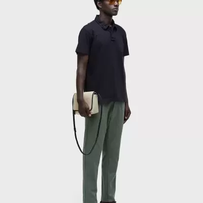 Pessoa com polo preta, calças verde-acinzentado, sapatos castanho escuro, óculos amarelos e bolsa bege com alça preta