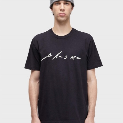 Pessoa com t-shirt preta com texto Alaska, calças pretas e gorro cinza