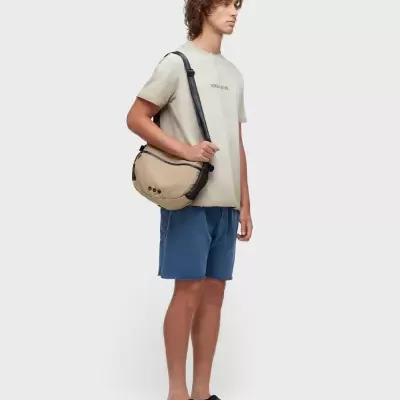 Homem com t-shirt bege, calções azuis, chinelos pretos e bolsa bege com correia preta ao ombro