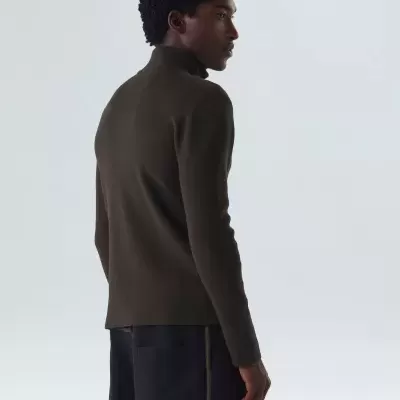 Homem com camisola castanha escura de gola alta e calças pretas em fundo branco.