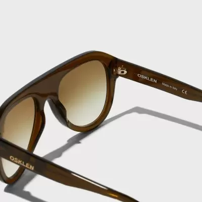 Óculos de sol castanhos translúcidos com lentes escuras degradé e texto nas hastes.