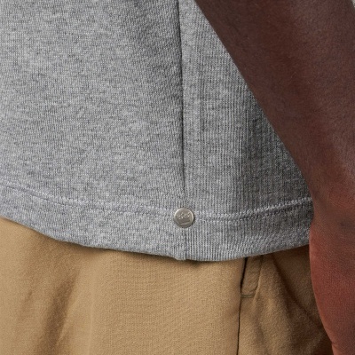 Detalhe de t-shirt cinza canelada com etiqueta metálica junto a calças bege e braço