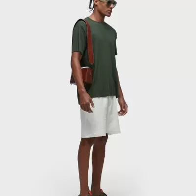 Homem com roupa casual verde e branca, bolsa e sandálias, em fundo branco.