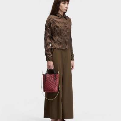 Mulher com camisa castanha e calças largas castanhas segurando mala vermelha com padrão de escamas