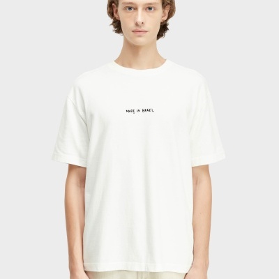 T-shirt branca com texto MADE IN BRAZIL preto ao centro, pessoa com calças bege, fundo cinza.
