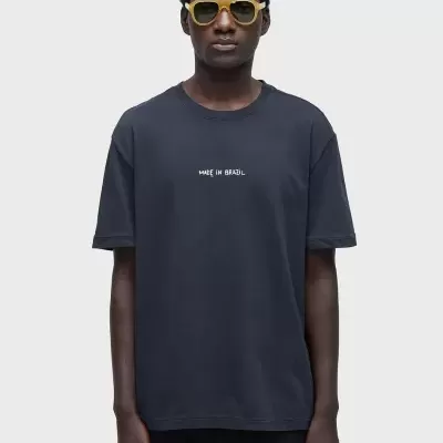 T-shirt preta com texto branco MADE IN BRAZIL e óculos amarelos num modelo contra fundo cinzento claro.