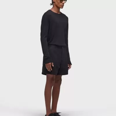 Homem com roupa preta, óculos escuros, calções e ténis, sobre fundo cinzento claro.