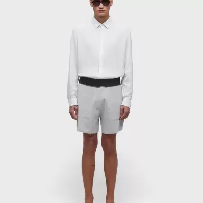 Homem com camisa branca, calções cinza, cinto preto, sandálias pretas e óculos escuros em fundo branco.