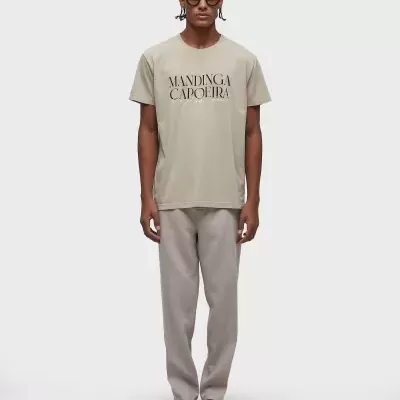 Homem com t-shirt bege com texto 'MANDINGA CAPOEIRA', calças cinza e chinelos