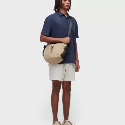 Homem jovem com camisa azul-marinho, calções bege, ténis bege e bolsa tiracolo bege com detalhes pretos e texto OSKLEN