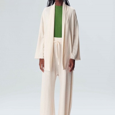 Mulher vestindo casaco creme, calças largas creme, t-shirt verde e ténis bege em fundo cinza claro.