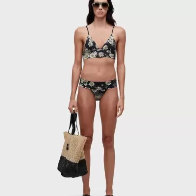 Mulher com bikini de flores margaridas em fundo preto, óculos de sol verdes, chinelos pretos e bolsa de mão bege e preta de malha