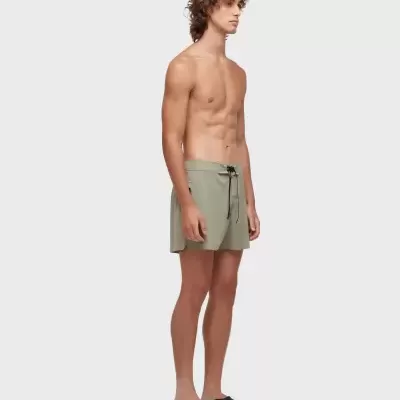 Homem de perfil com calções de banho verde-acinzentados e chinelos pretos