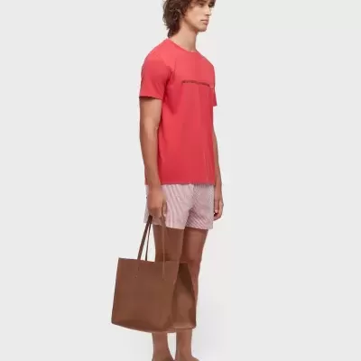 Homem com t-shirt vermelha, calções risca vermelha branca, sandálias bege e mala castanha de camurça.