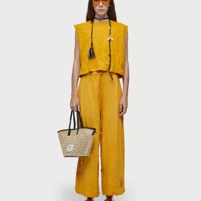 Conjunto amarelo com top e pantalonas, óculos amarelos, colar preto, bolsa de palha e sapatos castanhos.
