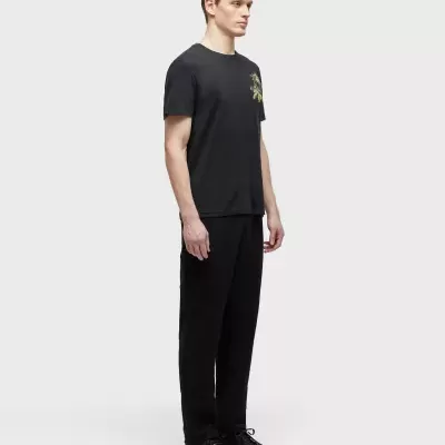 Homem de pé com t-shirt preta de manga curta com padrão verde no peito, calças e sapatos pretos, fundo branco.