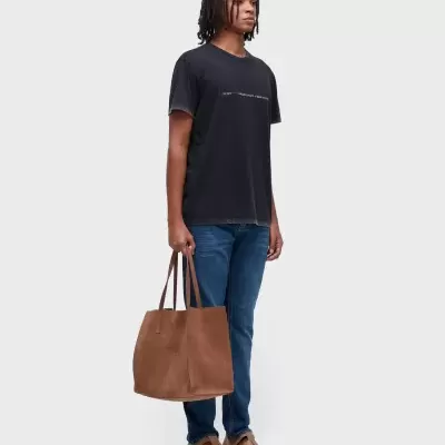 Homem com t-shirt preta, jeans azuis, sandálias castanhas e mala tote castanha