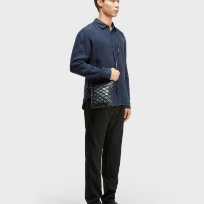 Homem com camisa azul escura, calças pretas, chinelos pretos texturizados e mala preta tiracolo.