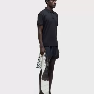 Homem com polo preta, calções pretos, sandálias pretas e lenço preto e branco abstrato num fundo branco.