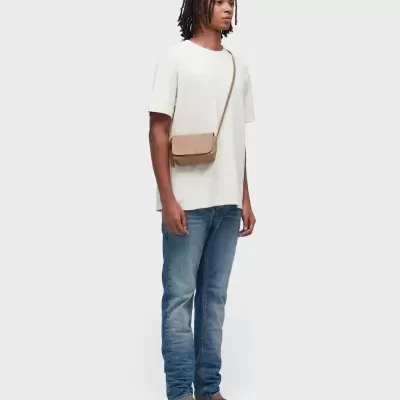 Pessoa vestindo t-shirt branca, jeans azuis, sandálias castanhas e bolsa bege cruzada.