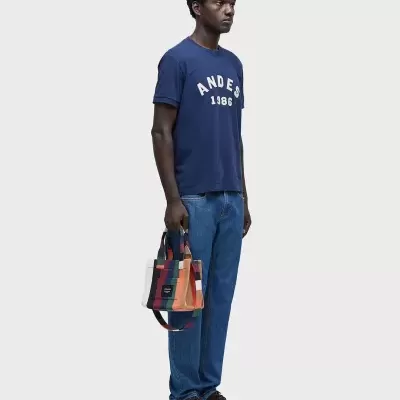 Homem com t-shirt azul, jeans, sapatos escuros e mala xadrez colorida