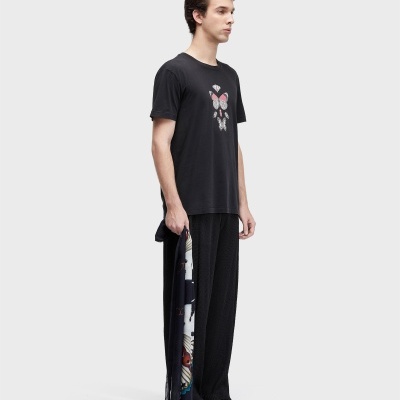 Homem de pé com t-shirt preta estampada e calças largas pretas em fundo branco