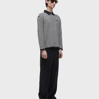 Homem com camisa às riscas preto e branco, calças pretas largas e sapatilhas bege em fundo cinzento