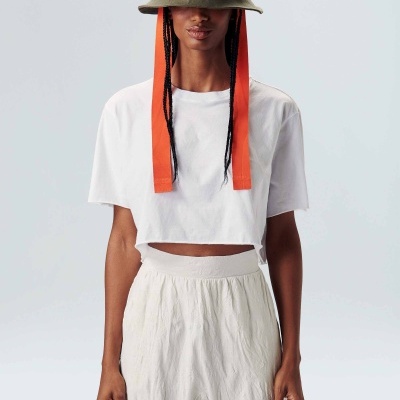 Mulher com chapéu verde com fitas vermelhas, t-shirt branca curta e calças largas brancas em fundo claro