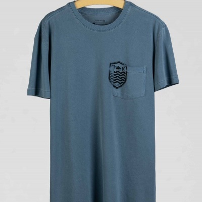 T-shirt azul com bolso e crachá na frente, pendurada num cabide de madeira