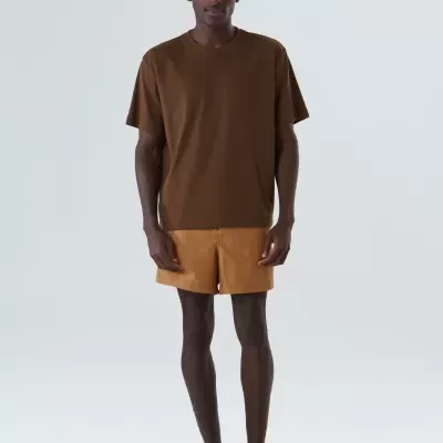 Homem com t-shirt castanha, calções cor de caramelo e ténis castanhos em fundo branco.