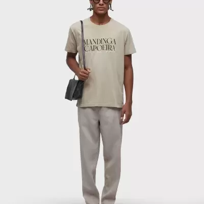 Homem com t-shirt bege com texto, calças cinza, sandálias, óculos de sol e mala preta em fundo branco