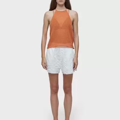 Mulher vestindo top laranja texturizado, calções brancos e sandálias negras, com óculos escuros em fundo cinza.